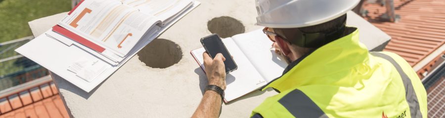 Urban roofer taking notes holding iPhone and writing in an urban location wearing hard hat and safety jacket, Fast Forward Commercial Excellence